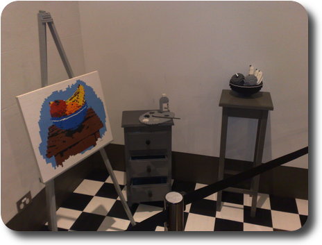 Artist's corner, with table and bowl of fruit, chest of drawers and painting on easel