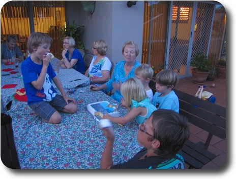 Child sitting on table eating cake, children and adults around table, some eating cake