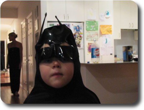 Little girl wearing a black mask and cape, standing in kitchen