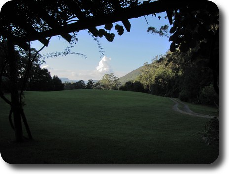 Towering cloud in blue sky, beyond lawn in shade, trellis in foreground