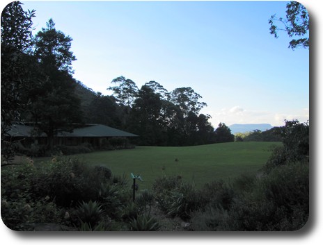 Garden in front, large lawn with long house to left, sun setting behind hill behind house