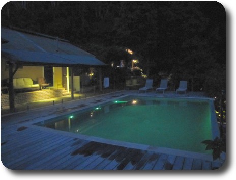 Night time photo of swimming pool, with house on one side, another back a little in the trees behind