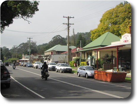 Two lane road, wiht old buildings on far side, parked cars and a couple of motorcycles riding along