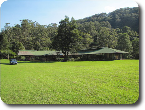 Large lawn, with car parked on it, back to long house, green roof and tree covered hills behind