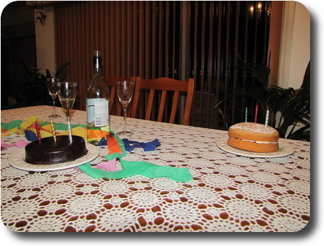 Two birthday cakes, each with 3 candles, on table with streamers and bottle of wine