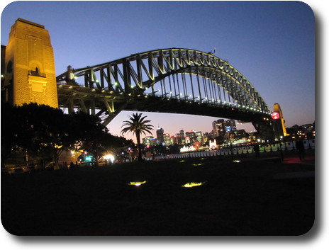 Dusk with illuminated arch bridge and high rise buildings visible across the water under the bridge