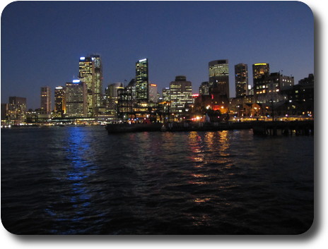 View over water to tall city buildings, with lights on, and darkening sky