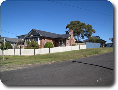 Brick house, grey roof and white fence on corner blokc; carport at rear