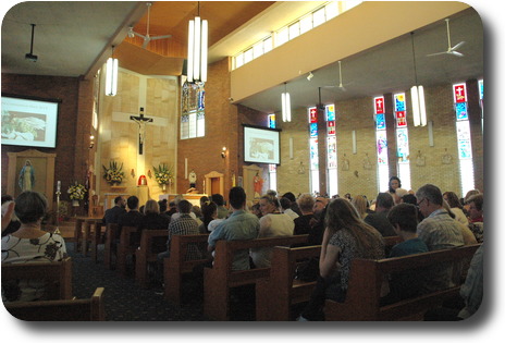 View from back left of church with seated congregation, high ceiling with windows below roof line