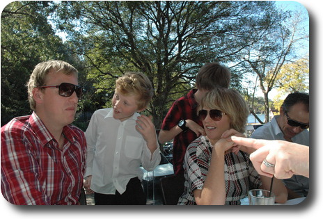 Three adults at table, two boys behind, one in conversation with an adult