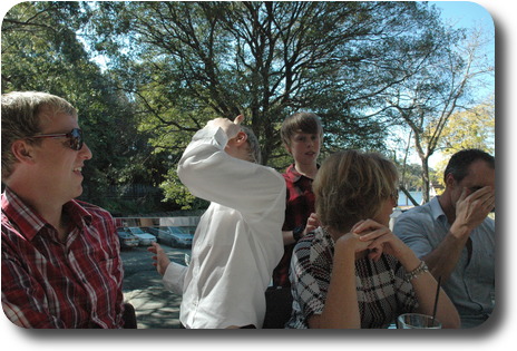 Three adults at table, two boys behind, one in conversation with an adult