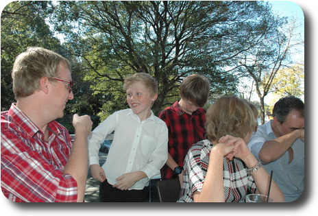 Three adults at table, two boys behind, one in conversation with an adult