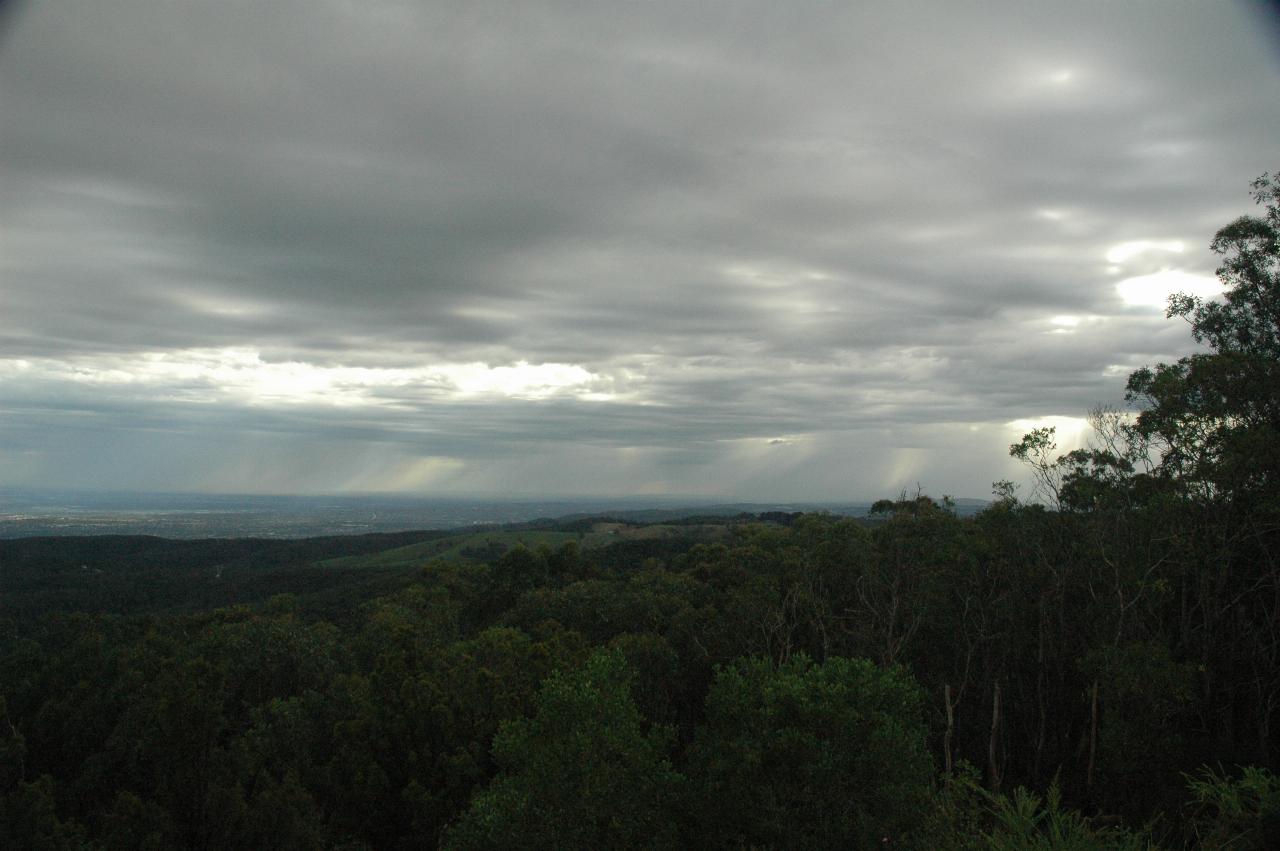 Foreground valley with trees, distant landscape with gray clouds and some rain falling from them