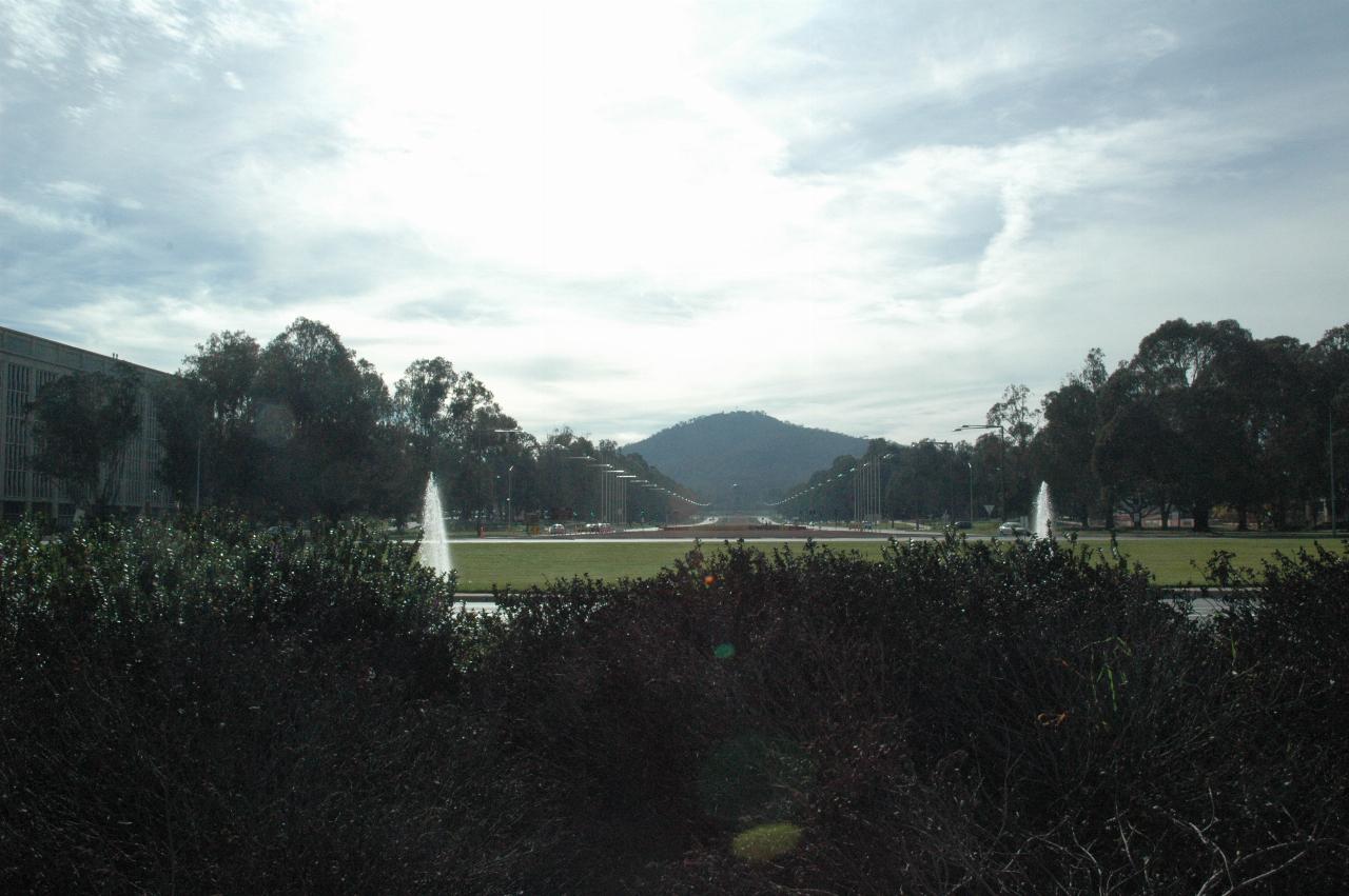 Looking over bushes, two water jets and road leading to distant dome shaped building and mountain behind
