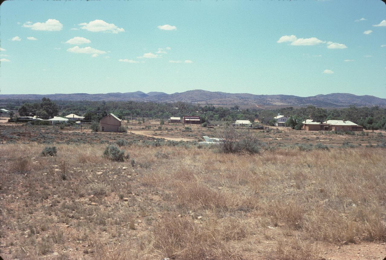 Few scattered buildings across dry country; barren hills in the distance