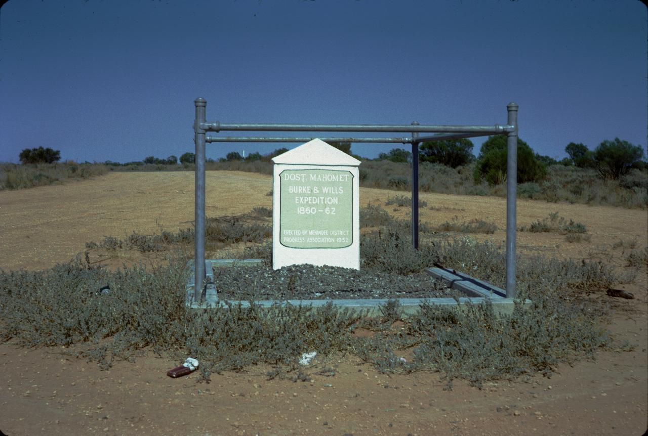Cement grave marker, protected by metal pipe railing at edge of dirt road